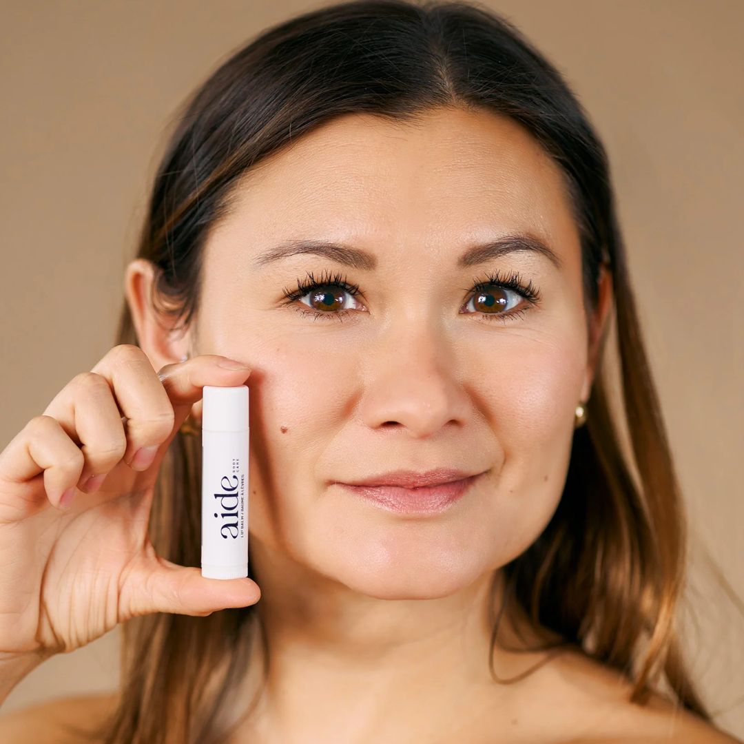 A person holding a  white tube of lip balm with the word "aide" printed in black on the label.