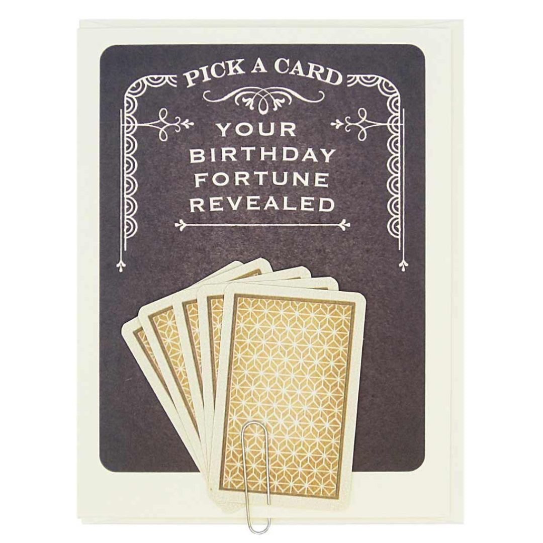 A dark grey greeting card with a white decorative border at the top and bottom. The top of the card reads "PICK A CARD" and "YOUR BIRTHDAY FORTUNE REVEALED." Below this, five fan-arranged cards with a gold and white patterned back are held by a paperclip. A second image displays five individual fortune cards, each resembling a playing card with suit symbols and text. The fortunes include messages like "YOUR LUCK IS ABOUT TO CHANGE," "SOME SECRETS ARE BEST KEPT SECRET. SORRY," and "THERE IS HIDDEN MONEY IN A