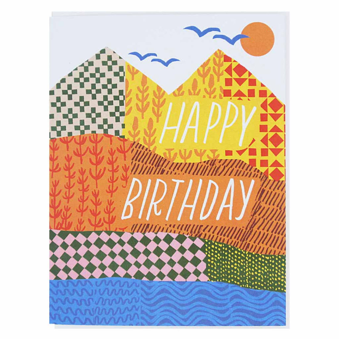 A colorful greeting card featuring a landscape design with mountains, a sun, birds, and a body of water. The mountains are depicted with various patterns and the words "HAPPY BIRTHDAY" are written in white across two of the mountain peaks.