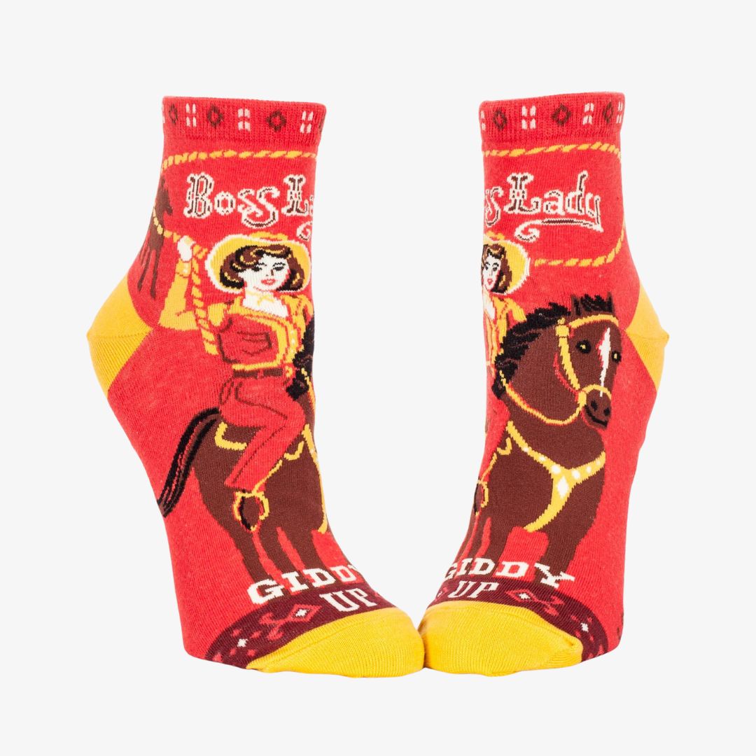 A pair of red ankle socks with yellow accents. One sock features a vintage-style illustration of a cowgirl riding a horse, with "Boss Lady" text above and "Giddy Up" below. The other sock shows the back of the cowgirl and horse with the "Giddy Up" text.