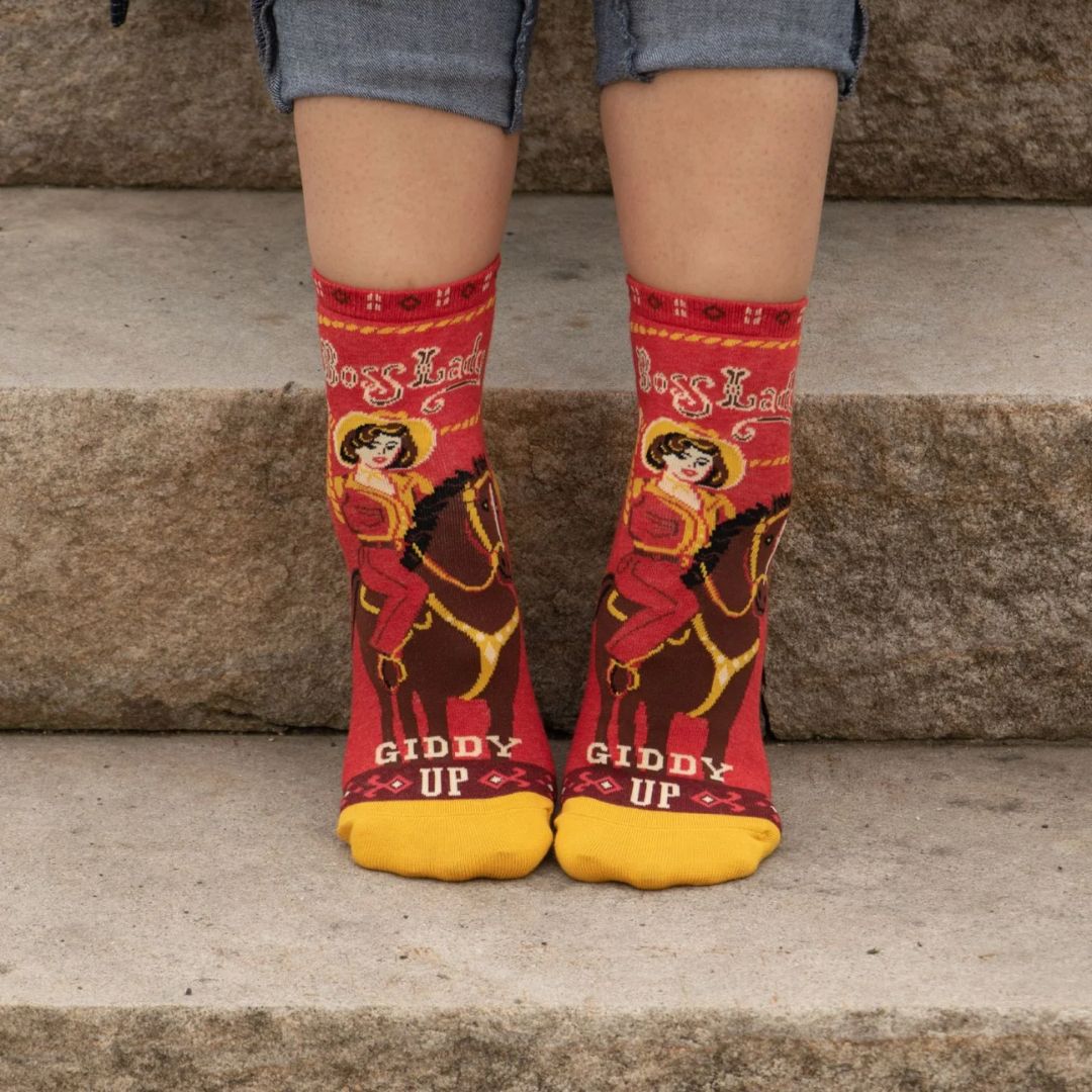 A pair of red ankle socks with yellow accents. One sock features a vintage-style illustration of a cowgirl riding a horse, with "Boss Lady" text above and "Giddy Up" below. The other sock shows the back of the cowgirl and horse with the "Giddy Up" text.