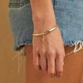 A set of two gold bracelets, one layered over the other, with a lightning bolt charm hanging from the bottom. The bracelets are displayed on a pink card with the brand name "Foxy Originals."