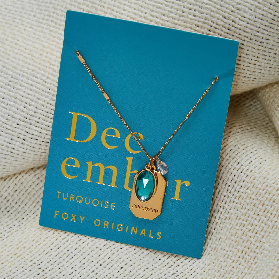 A gold necklace with a turquoise stone pendant. The pendant also has the word "FRIENDSHIP" engraved on it. The necklace is displayed on a blue card with the month "December" and the brand name "Foxy Originals."
