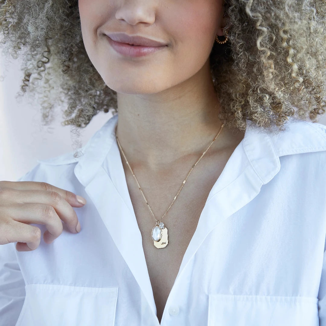 A gold necklace with a pearl stone pendant. The pendant also has the word "LOVE" engraved on it. The necklace is displayed on a blue card with the month "June" and the brand name "Foxy Originals."