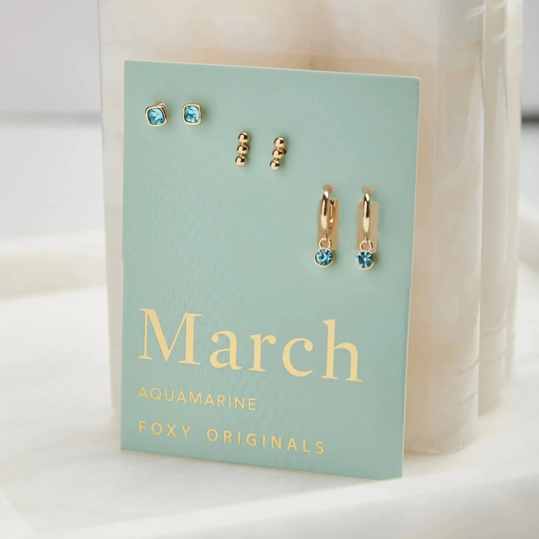 A set of three pairs of aquamarine earrings displayed on a light blue card. The set includes a pair of stud earrings, a pair of bar earrings with small aquamarine beads, and a pair of hoop earrings with dangling aquamarine stones. The card has the month "March" and the brand name "Foxy Originals."