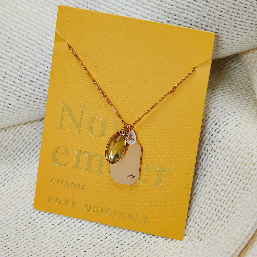 A gold necklace with a citrine stone pendant. The pendant also has the word "JOY" engraved on it. The necklace is displayed on a yellow card with the month "November" and the brand name "Foxy Originals."