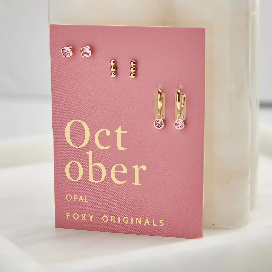 A set of three pairs of opal earrings displayed on a pink card. The set includes a pair of stud earrings, a pair of bar earrings with small opal beads, and a pair of hoop earrings with dangling opal stones. The card has the month "October" and the brand name "Foxy Originals."