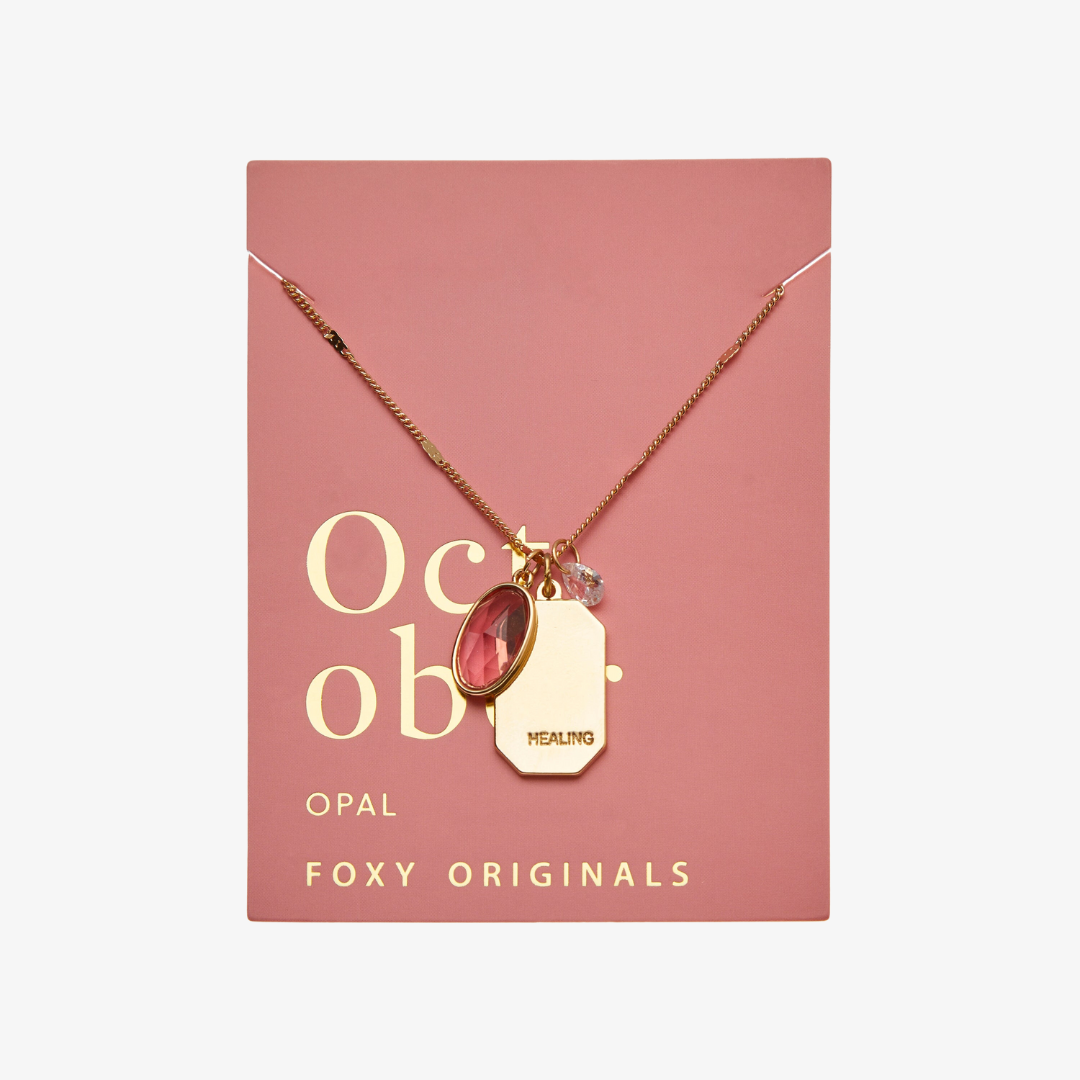 A gold necklace with an opal stone pendant. The pendant also has the word "HEALING" engraved on it. The necklace is displayed on a pink card with the month "October" and the brand name "Foxy Originals."