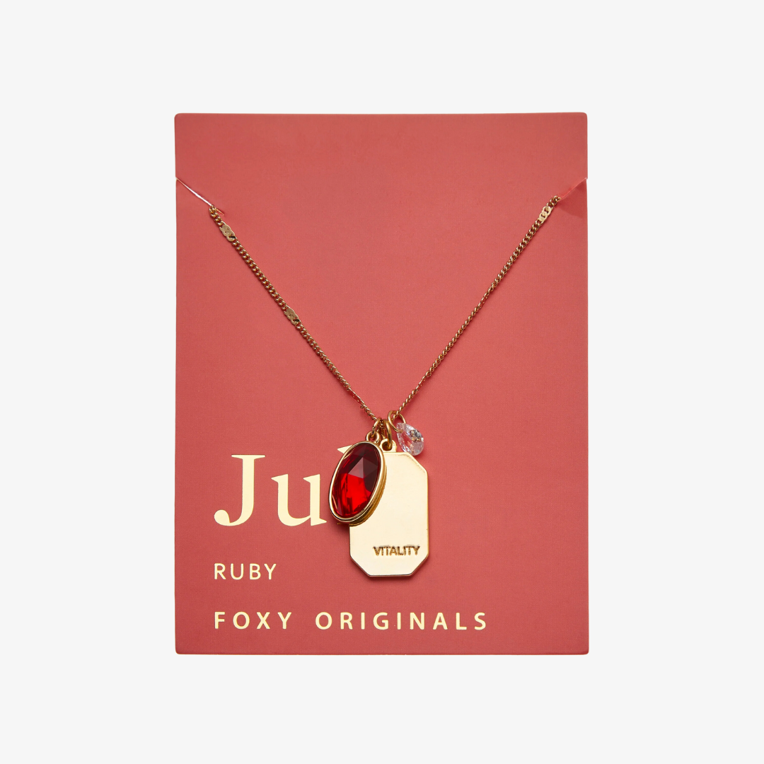 A gold necklace with a ruby stone pendant. The pendant also has the word "VITALITY" engraved on it. The necklace is displayed on a red card with the month "July" and the brand name "Foxy Originals."