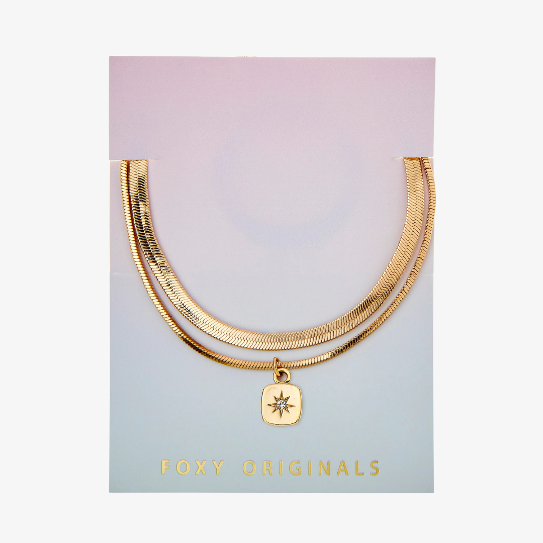 A set of two gold bracelets, one layered over the other, with a star charm hanging from the bottom. The bracelets are displayed on a pink card with the brand name "Foxy Originals."