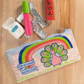A rectangular light blue pencil case with a zipper, featuring a whimsical illustration of a pink peacock with an outstretched rainbow tail feathers. Above the peacock is a rainbow arc and a banner with the text "I'm so casual and laid back".