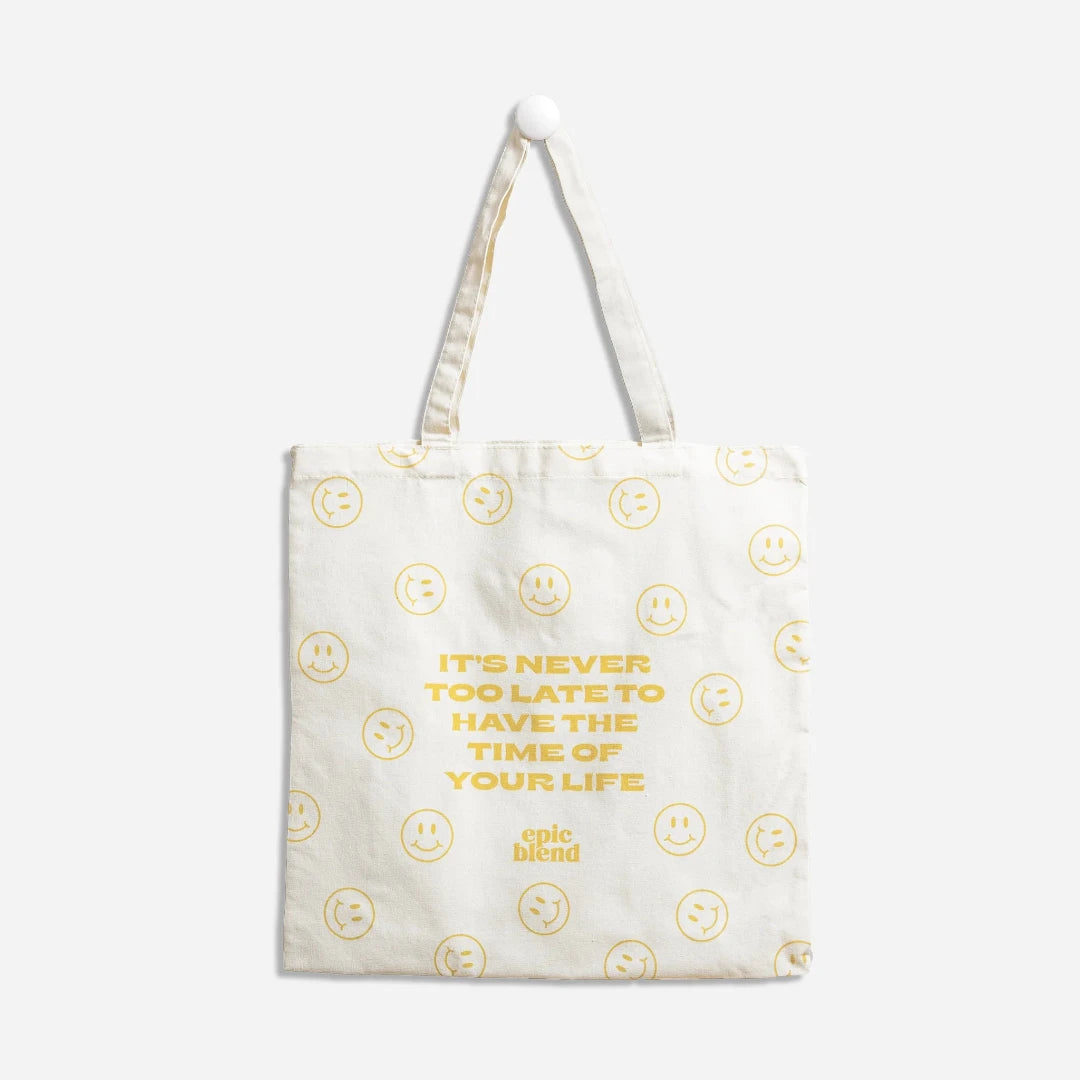 A white tote bag with a yellow smiley face design. The text "It's never too late to have the time of your life" is printed in yellow on the bag, surrounded by smiley faces. The "Epic Blend" logo is also printed in yellow on the bag.