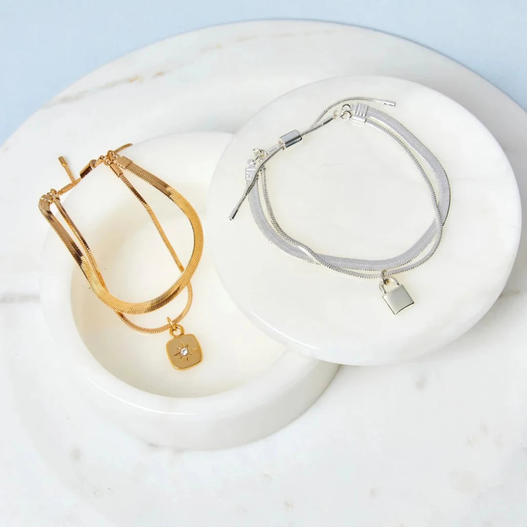A set of two gold and silver bracelets, one layered over the other, with a star charm hanging from the bottom. The bracelets are displayed on a pink card with the brand name "Foxy Originals."