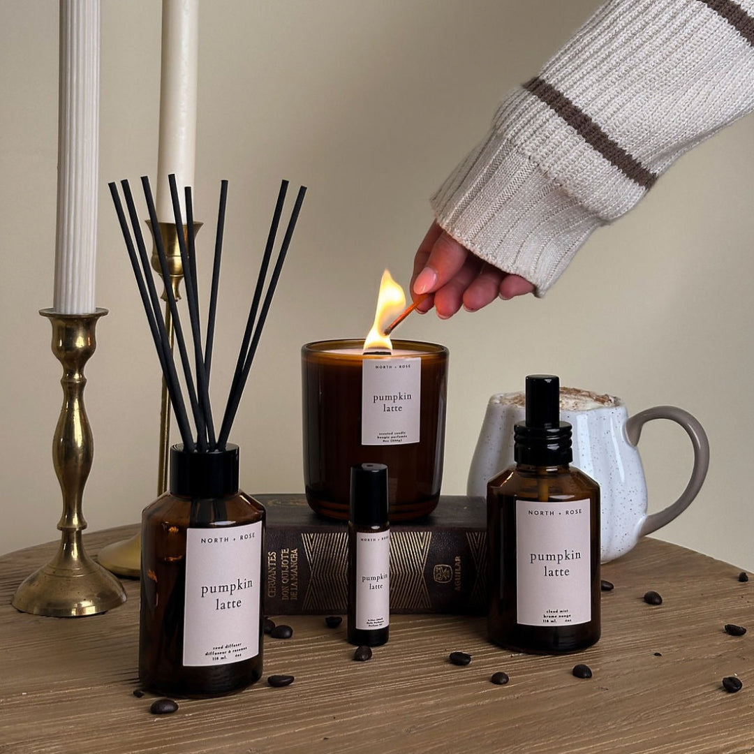 Person lighting a candle with diffusers and bottles labeled 'pumpkin latte' on a wooden surface.
