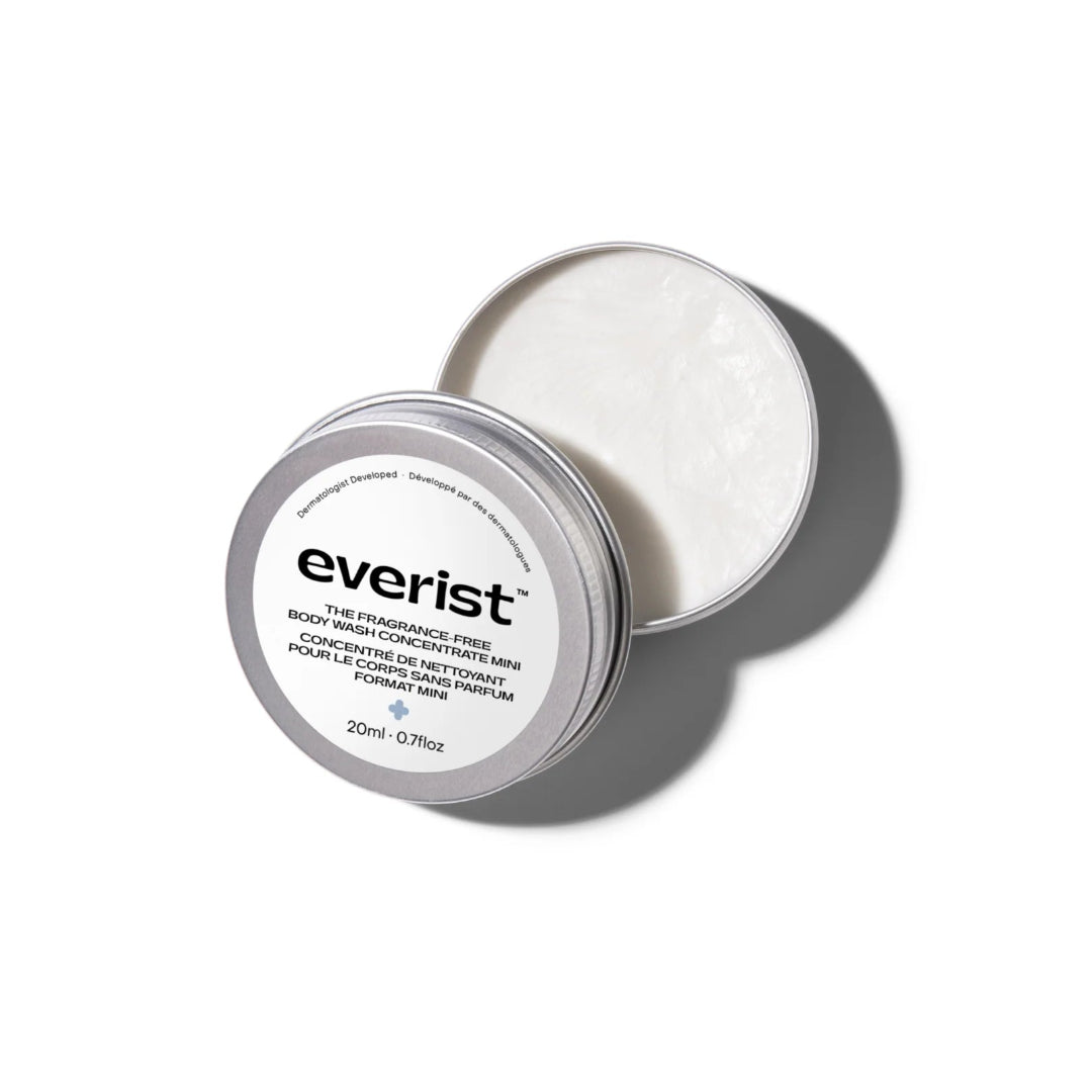 A small, open, silver metal tin of Everist's "The Fragrance-Free Body Wash Concentrate Travel Mini," revealing a white solid product inside. The tin's lid is propped against the base, and text on the side indicates it's dermatologist-developed and contains 20ml of product.