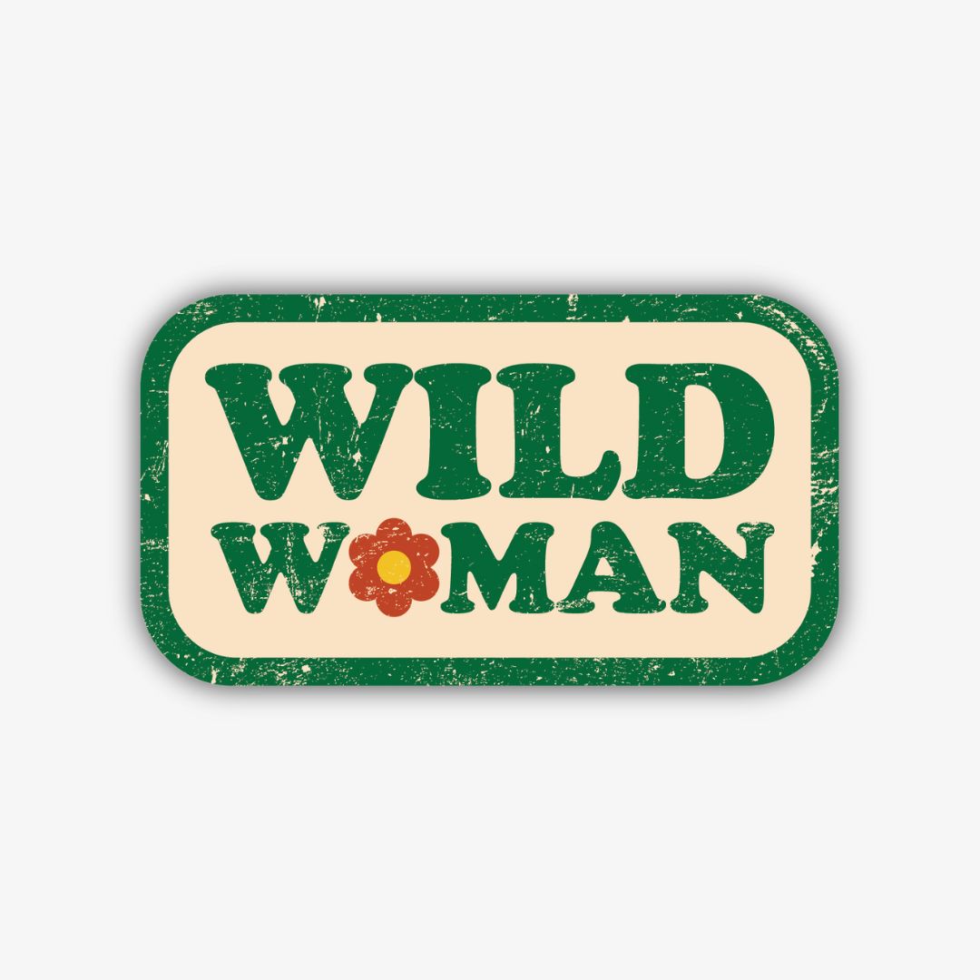A retro-style rectangular sticker with rounded corners, featuring a distressed light cream background and a dark green border. The words "WILD" and "WOMAN" are stacked in bold, distressed green lettering, with a reddish-orange flower replacing the 'O' in "WOMAN".